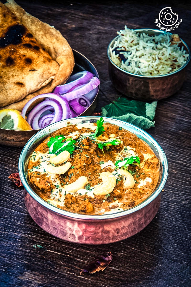 kaju curry garnsihed with cashews and cilanrto served in a metal bowl with some naan and onions in the background