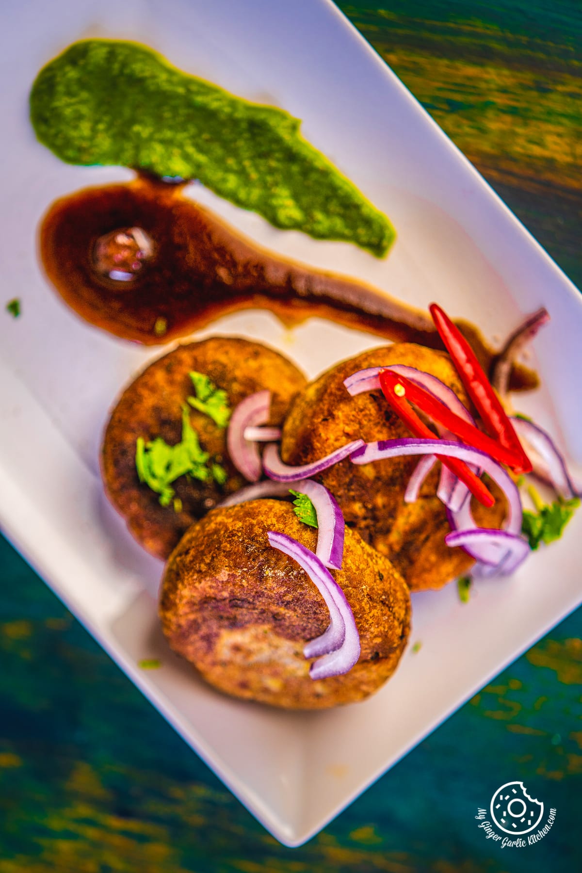 overhead shot of three aloo tikki served in a white plate with green coriander chutney and tamarind chutney
