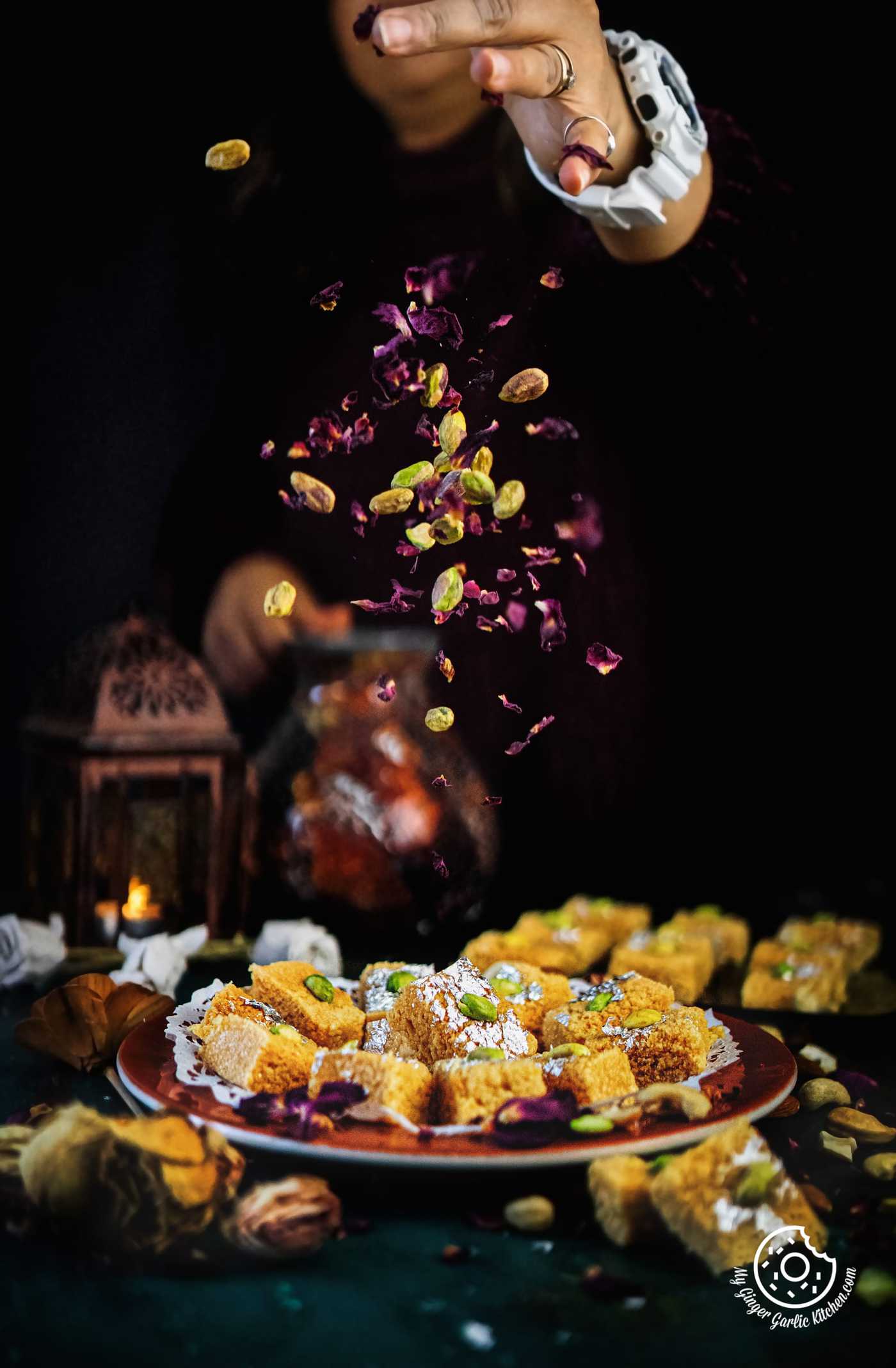 a feamle sprinkling dried rose petals and pisatchios over indian milk cake served in a brown plate