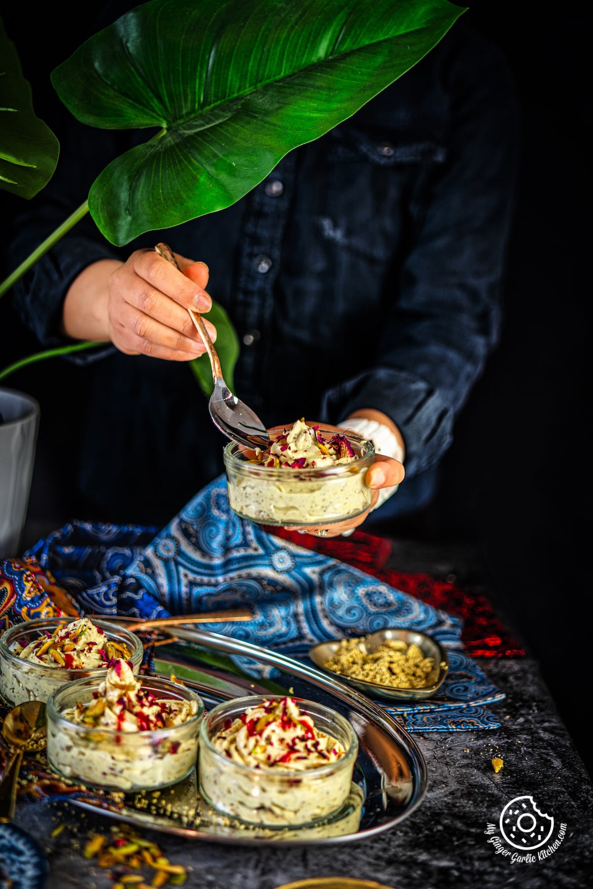 a female holding transparent bowl of thandai shrikhand and three other bowls in the frame