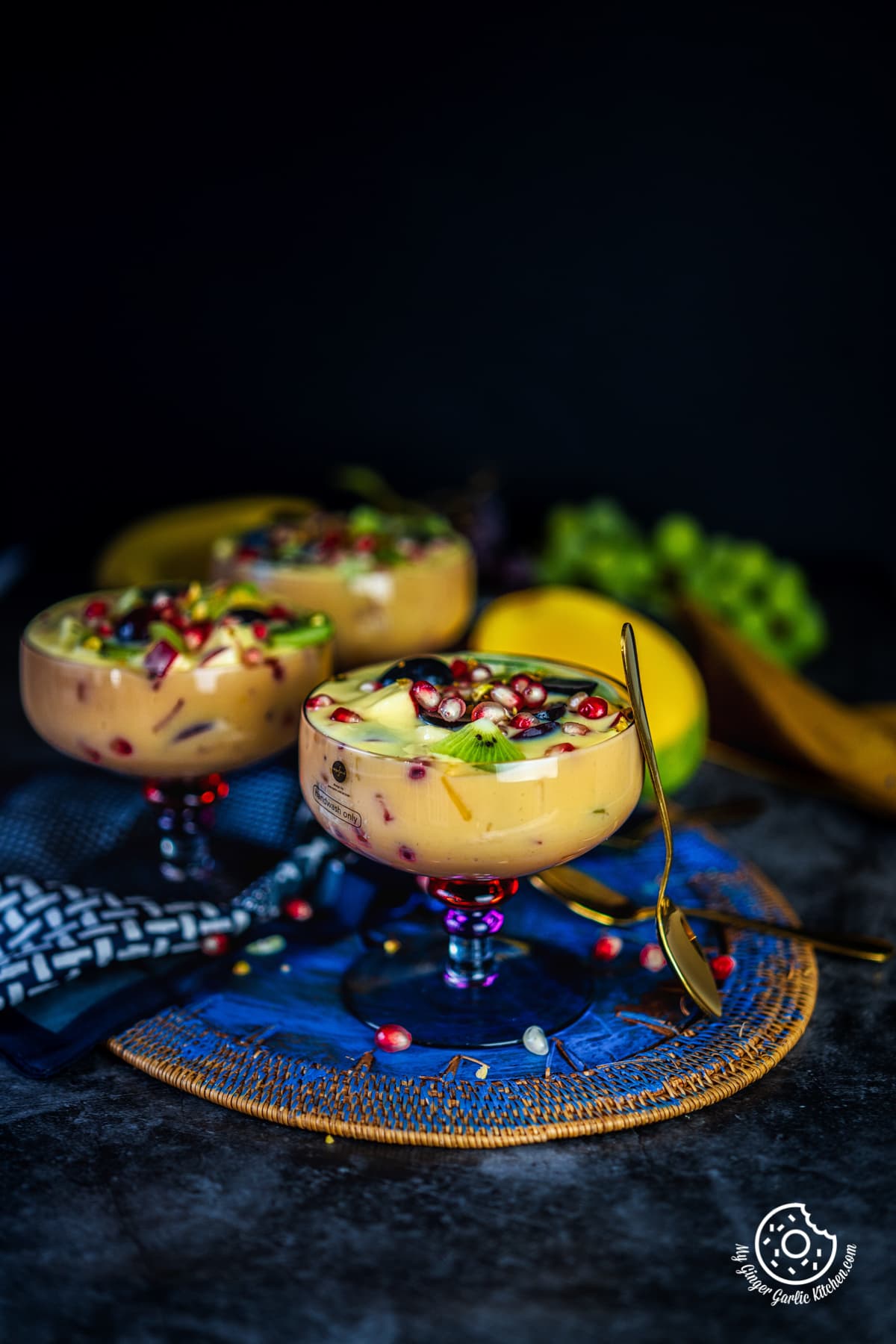 shot of fruit custard in a purple transparent bowl along with a golden spoon and two bowls in the background