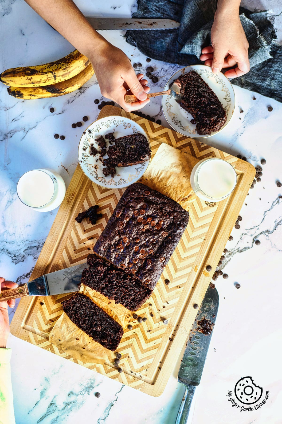 overhead shot of chocolate banana bread loaf on a wooden board and hands holding a white plate with bread slice
