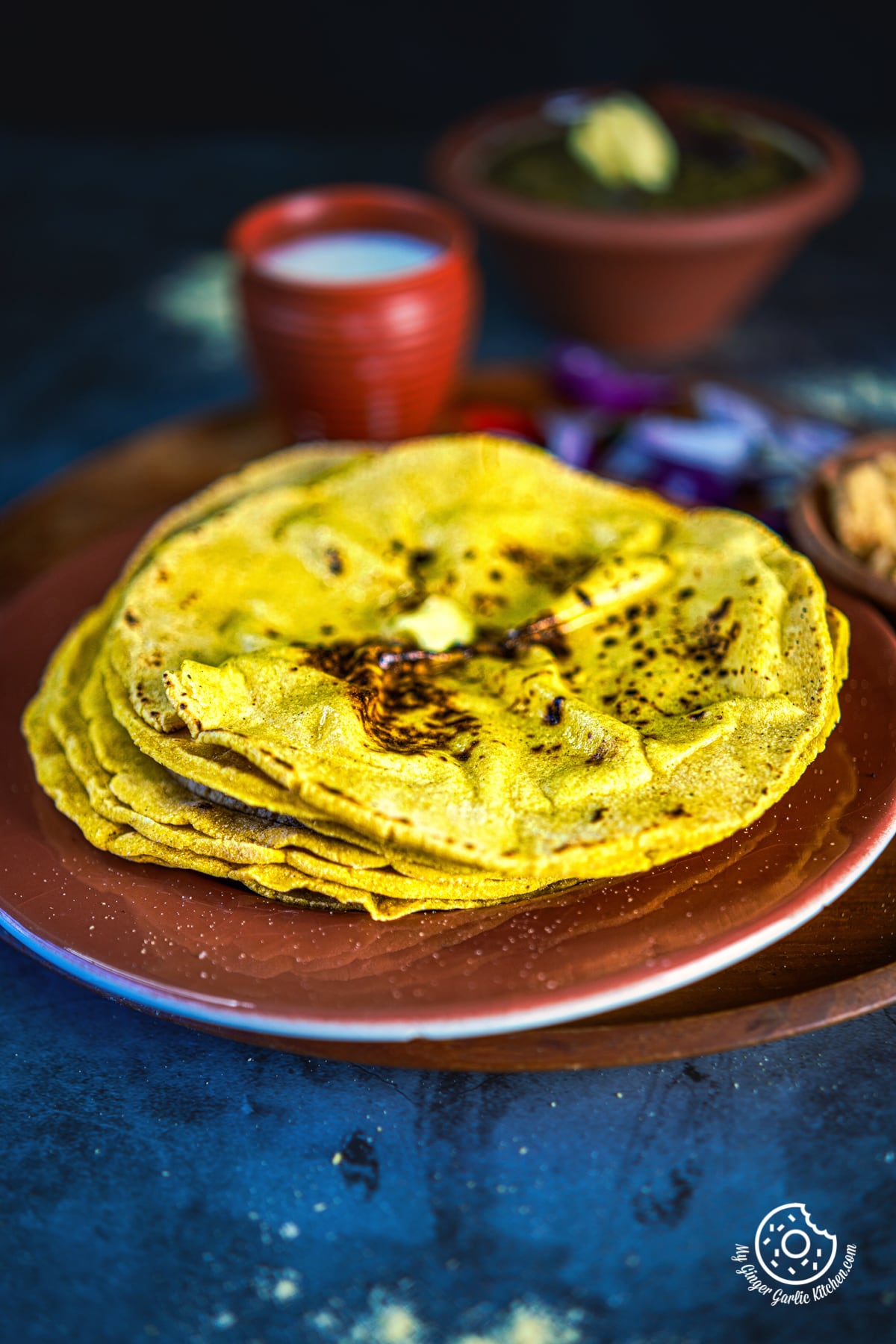 five makki ki roti topped with butter stacked in a brown plate with saag and buttermilk in the background