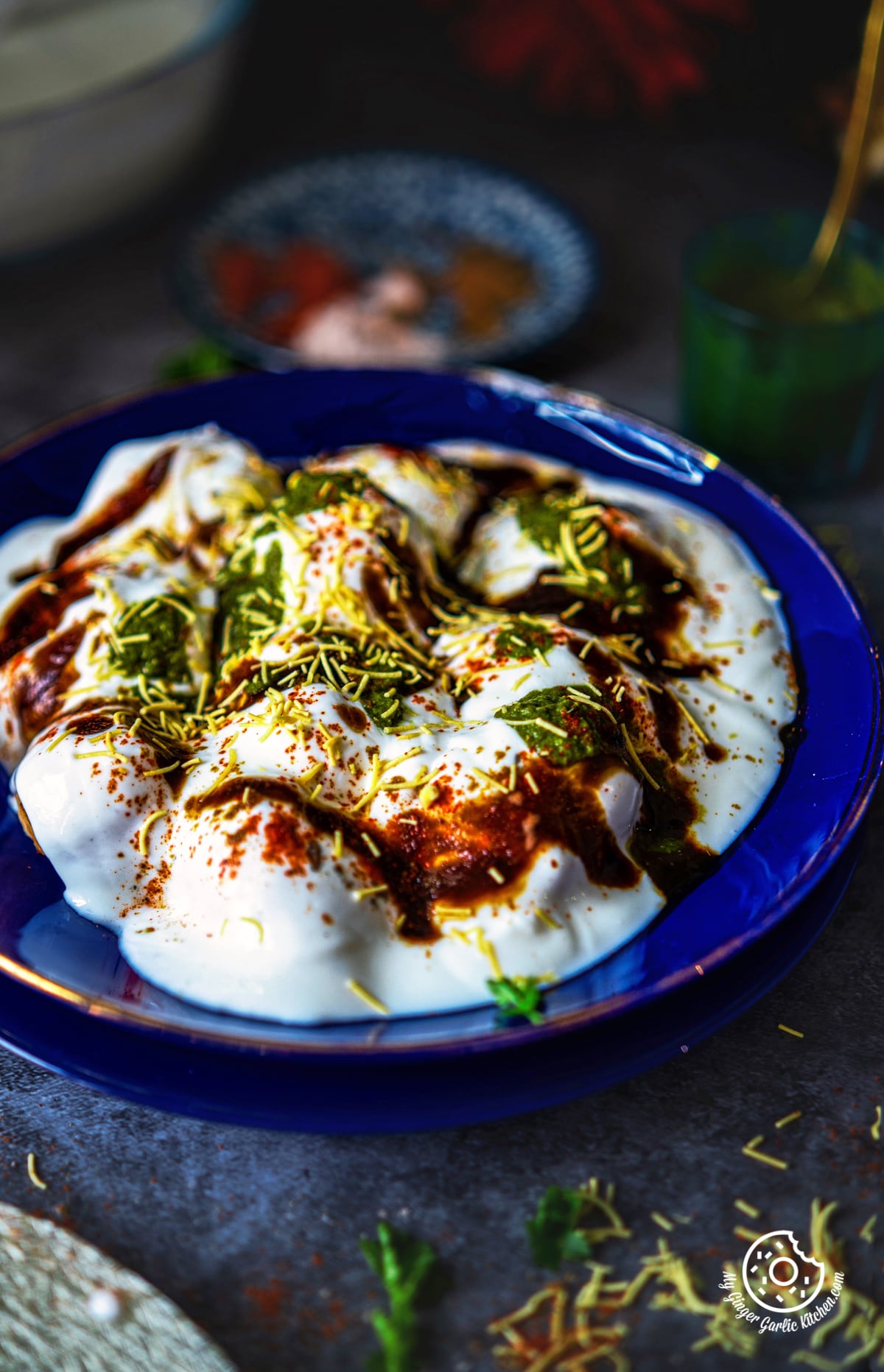 a closeup shot of dahi vada topped with chutneys and sev in a blue glass plate