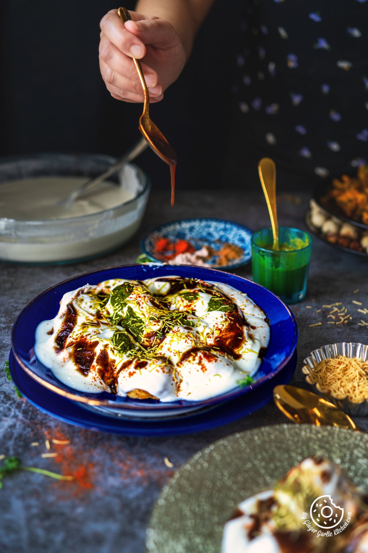 a hand pouring tamarind chutney over a dahi vada plate topped with chutneys and sev
