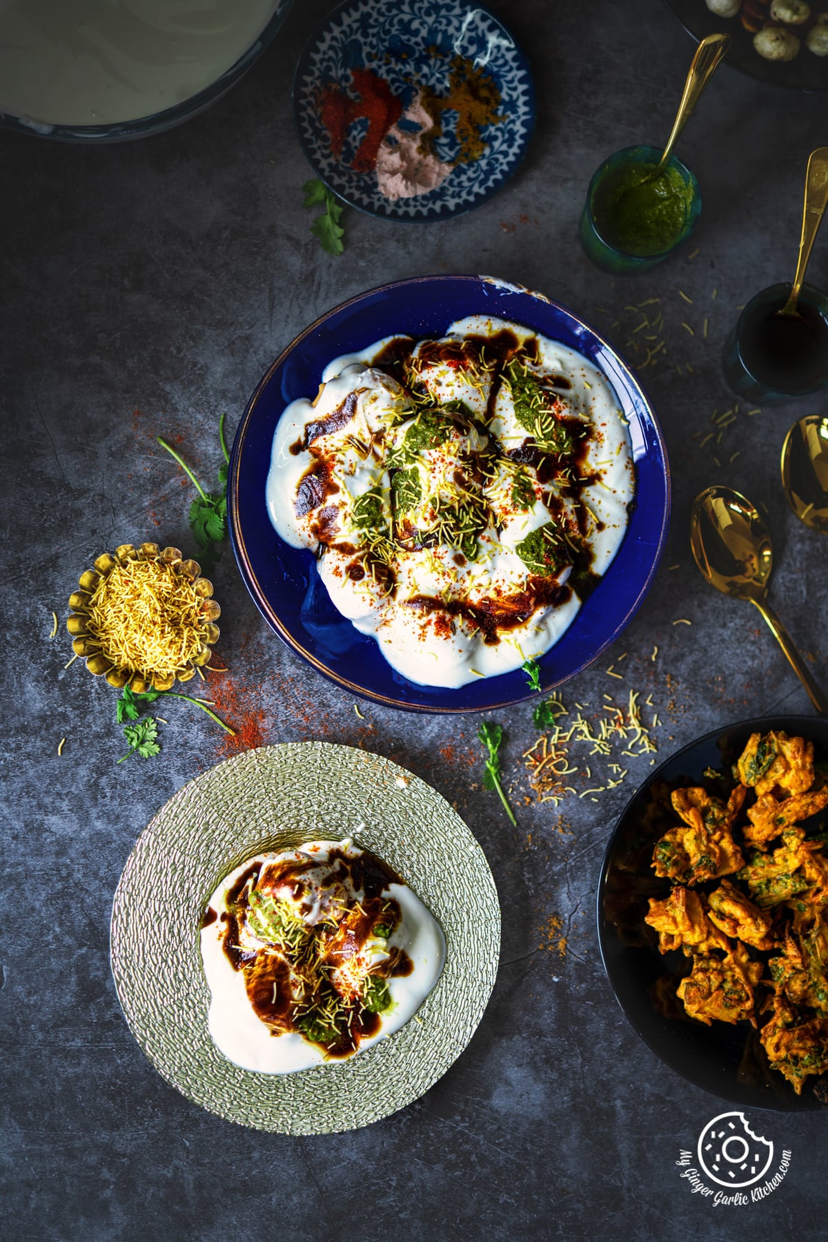 overhead shot of dahi vada in a blue glass plate topped with chutneys and sev with a silver plate filled with dahi on the side