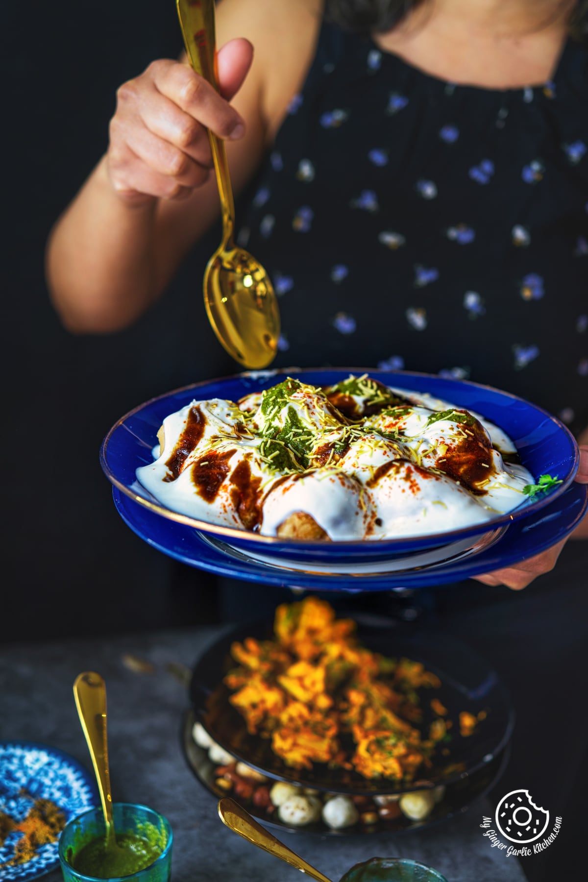a female holding a golden spoon in one hand and blue glass plate filled dahi vada topped with chutneys and sev with other hand