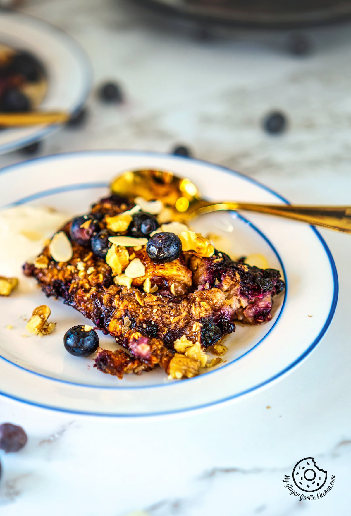 a closeup shot of a a white plate with a slice of banana blueberry baked oatmeal garnished with walnuts, almonds, yogurt, and blueberries