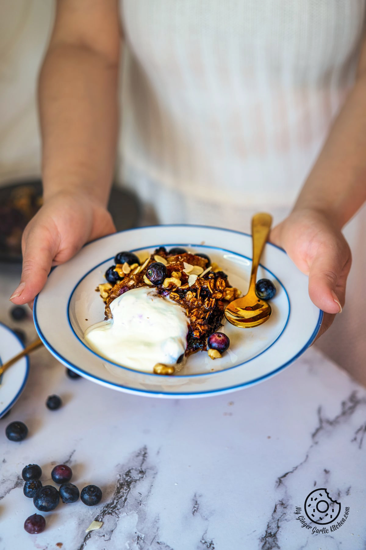 a female holding a white plate with a slice of banana baked oatmeal, walnuts, almonds, yogurt, and blueberries 