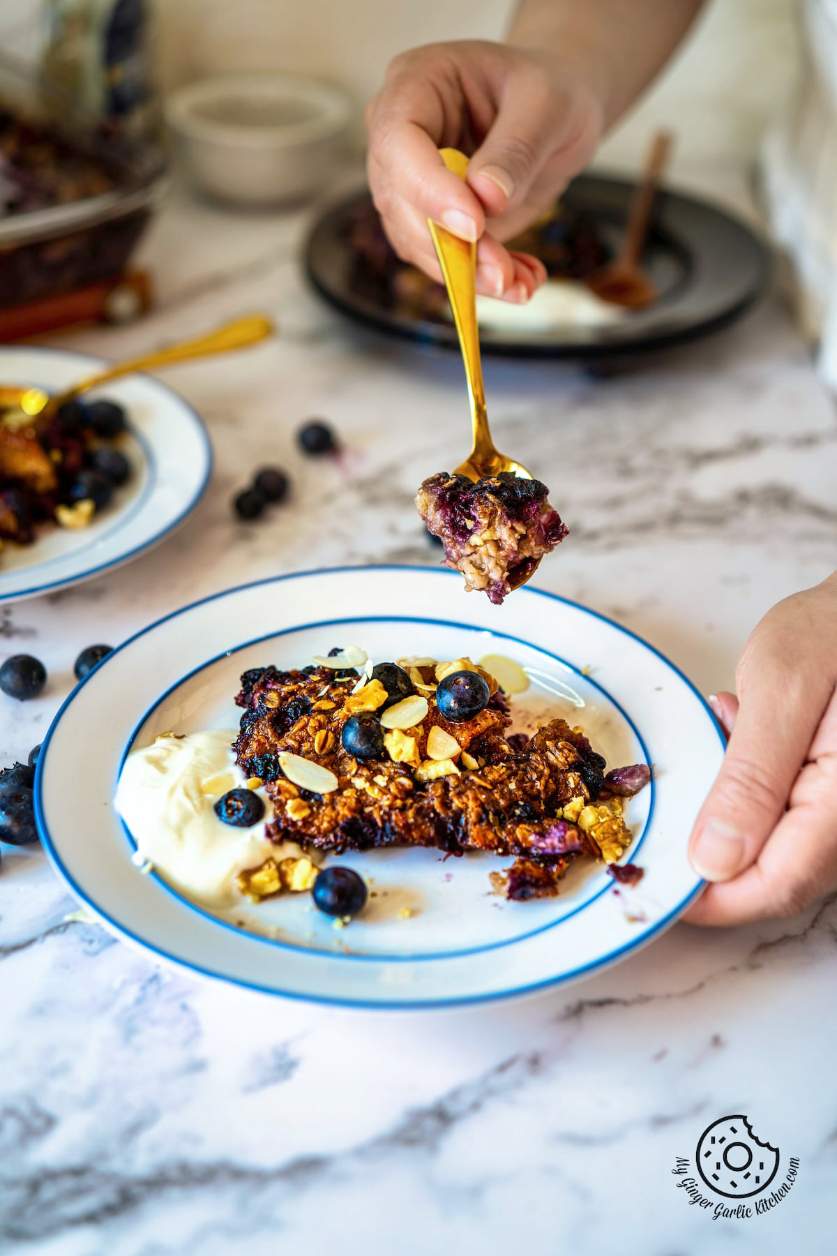 A golden spoon  filled with blueberry baked oats being lifted out of a white plate with a slice of baked oats garnished with blueberries