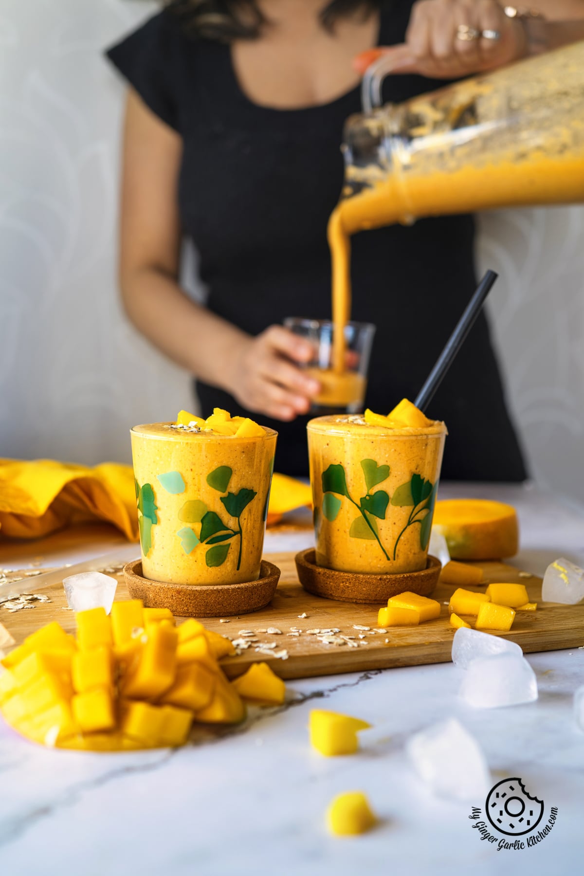 mango oats smoothie in two glasses with a female pouring smoothie in the background
