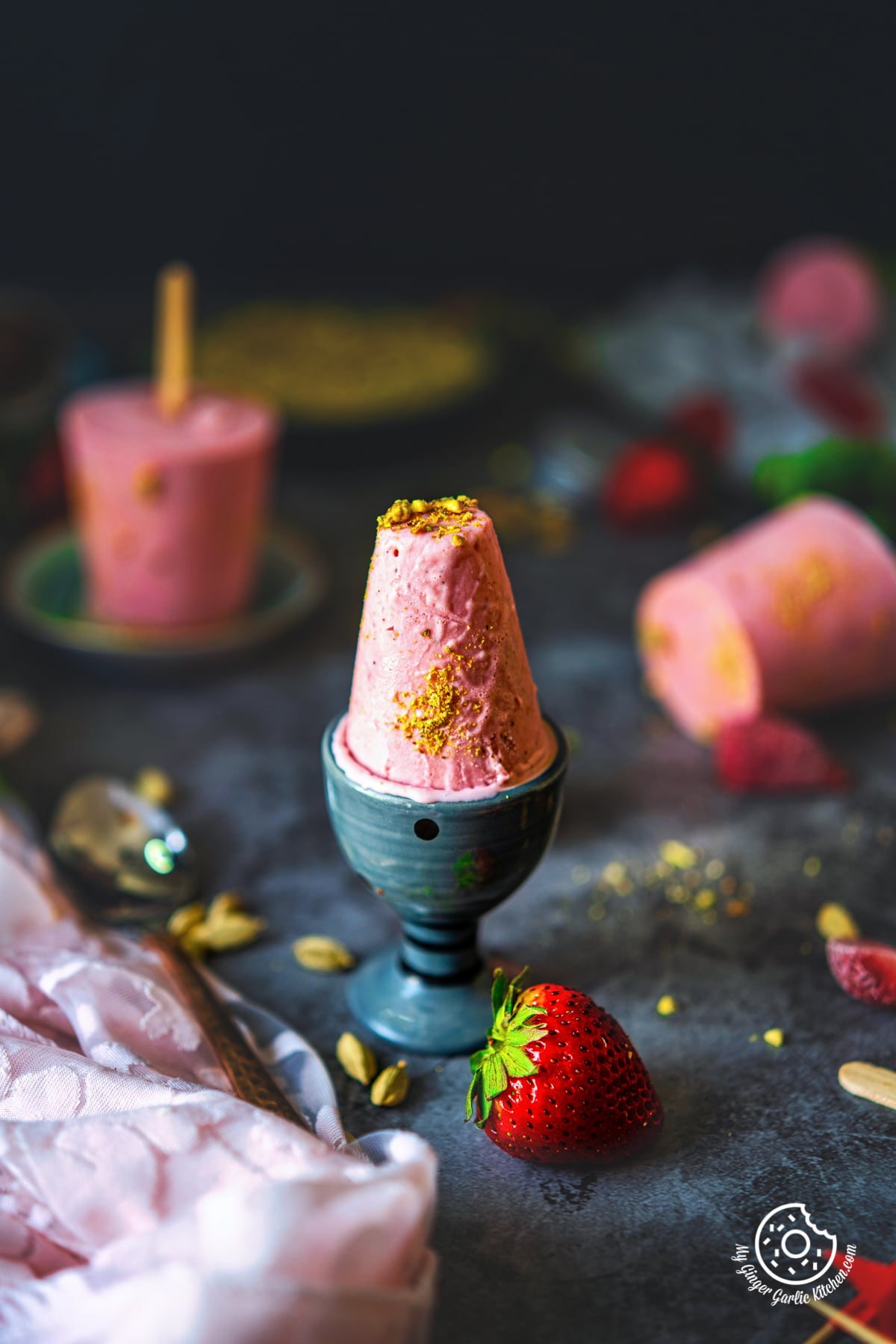 one strawberry kulfi in a small ceramic glass, with wo strawberry kulfis in the background