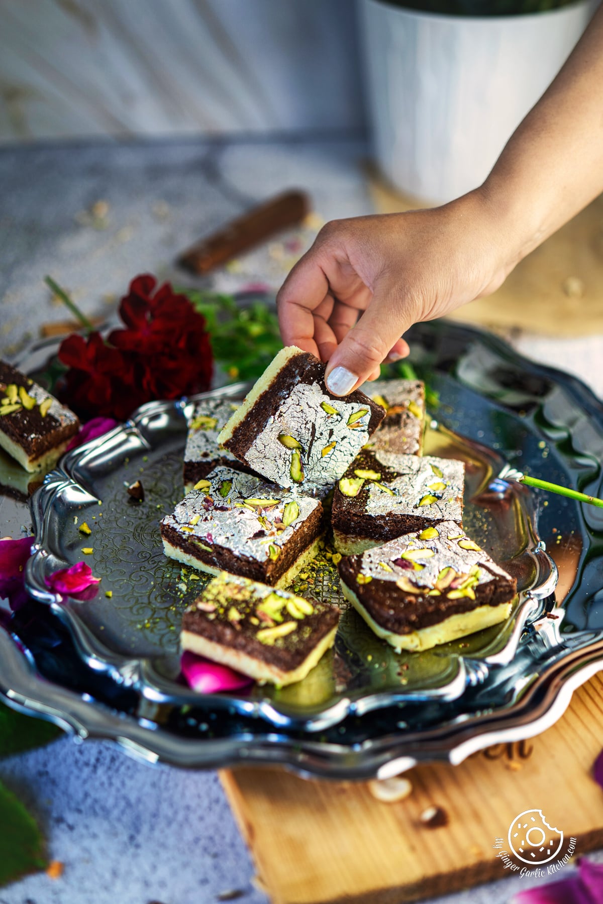 a hand holding chocolate burfi kept on a metal plate with flowers on the side