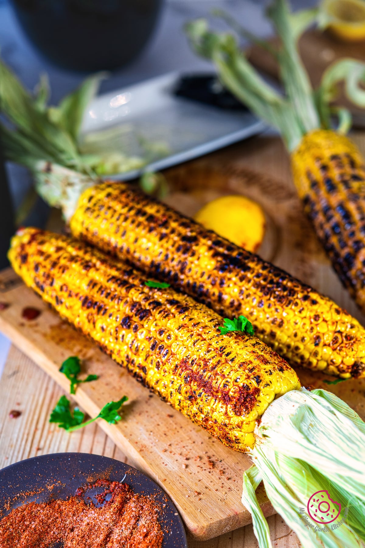 closeup photo of 2 indian roasted corn on the cobs with a spice bowl and lemon slice