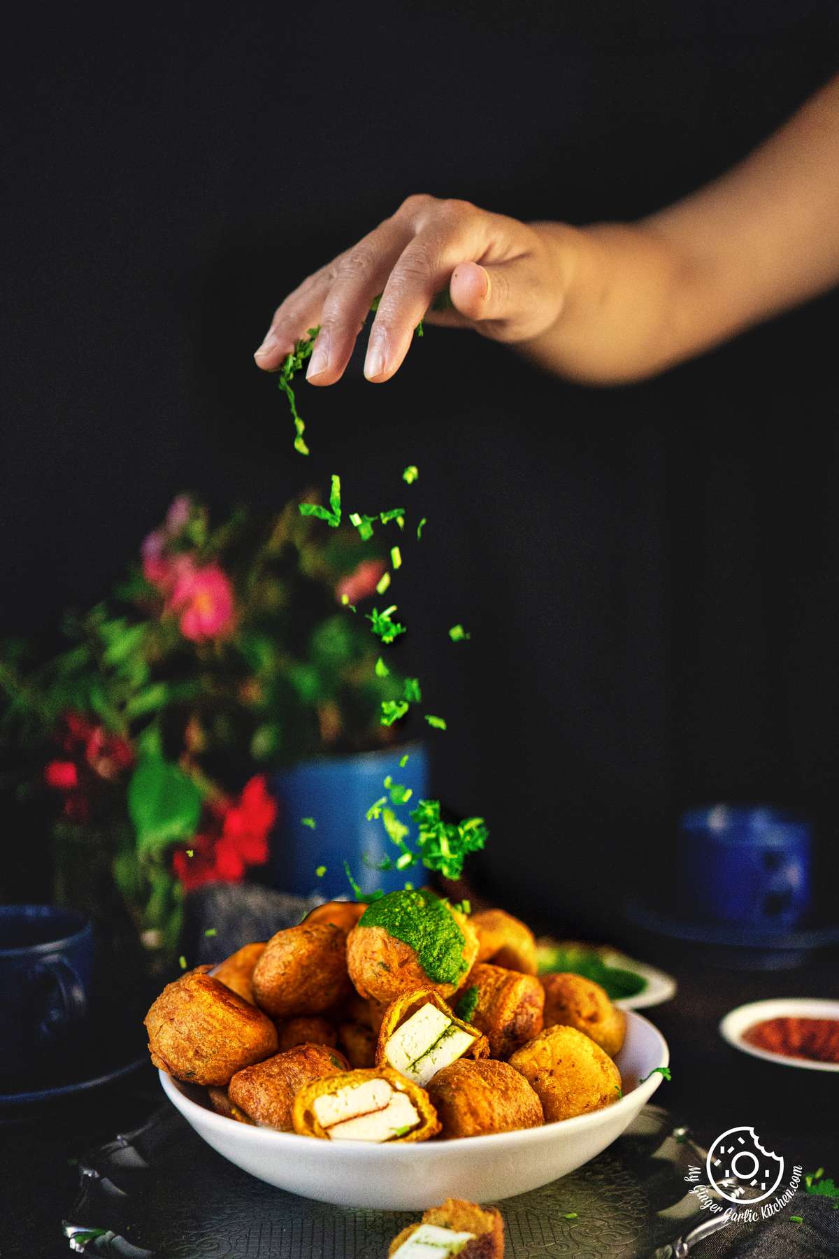 a hand sprinkling coriander leaves over a paneer pakora bowl