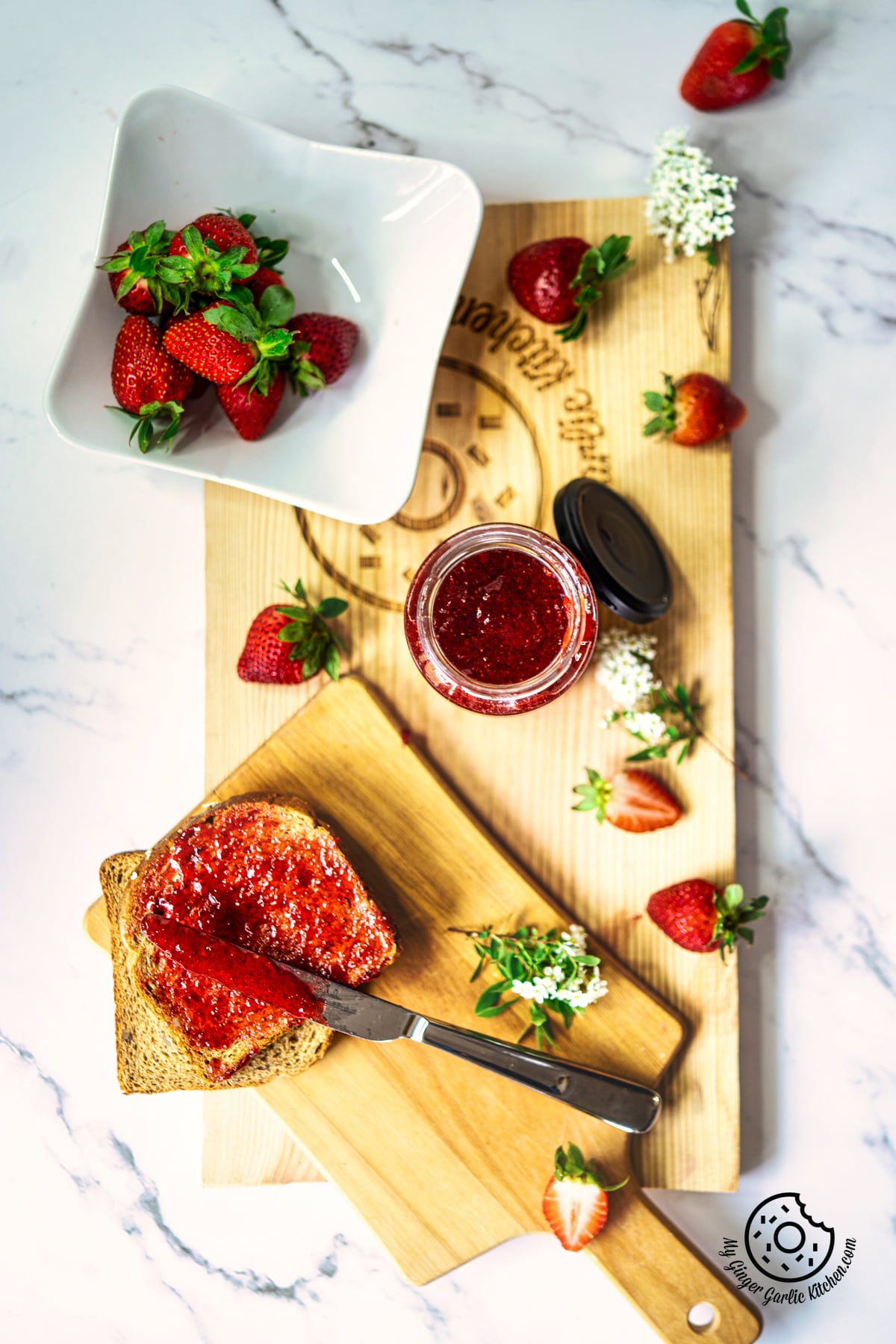 overhead shot of strawberry jam in a glass jar sitting on a wooden board plate with a bread slice and strawberries on the side