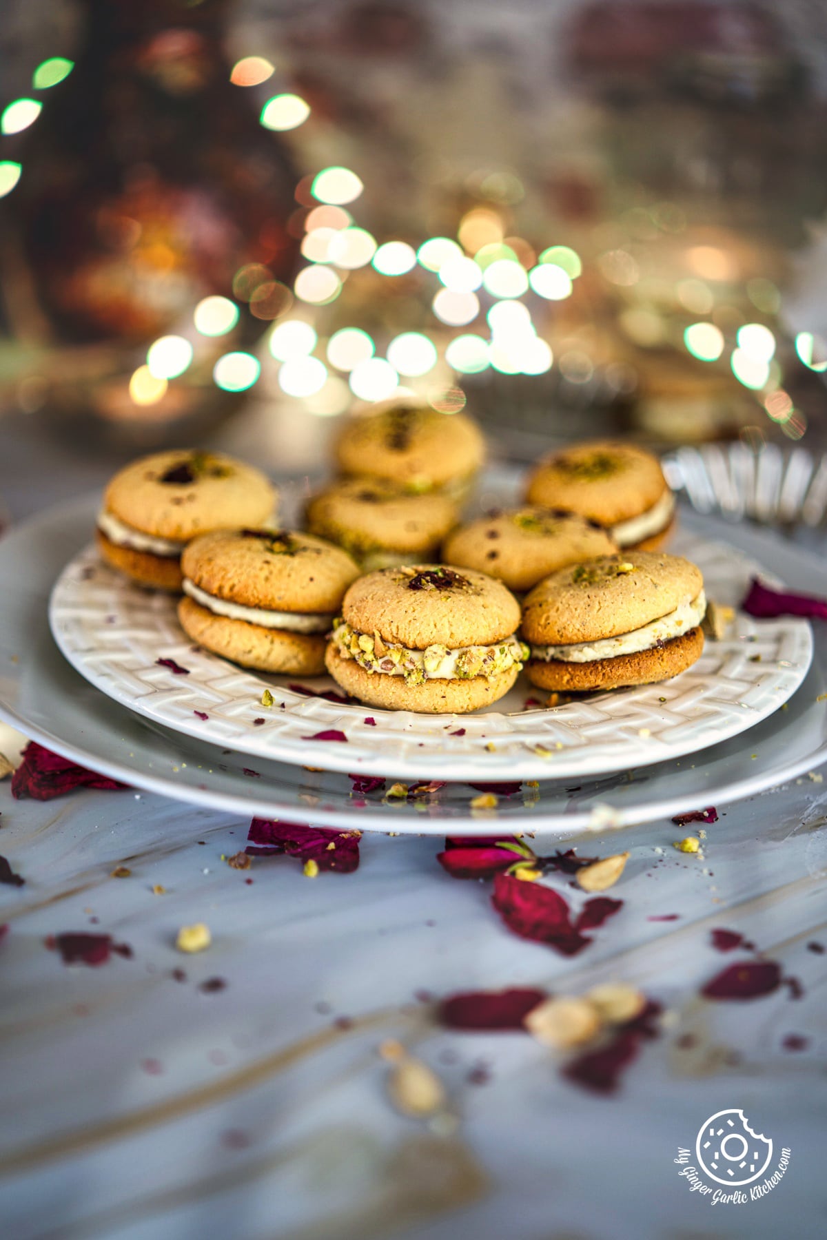 8 gulab jamun cookies served in a in a white plate with some dried rose petals on the side and bouquet light in the background