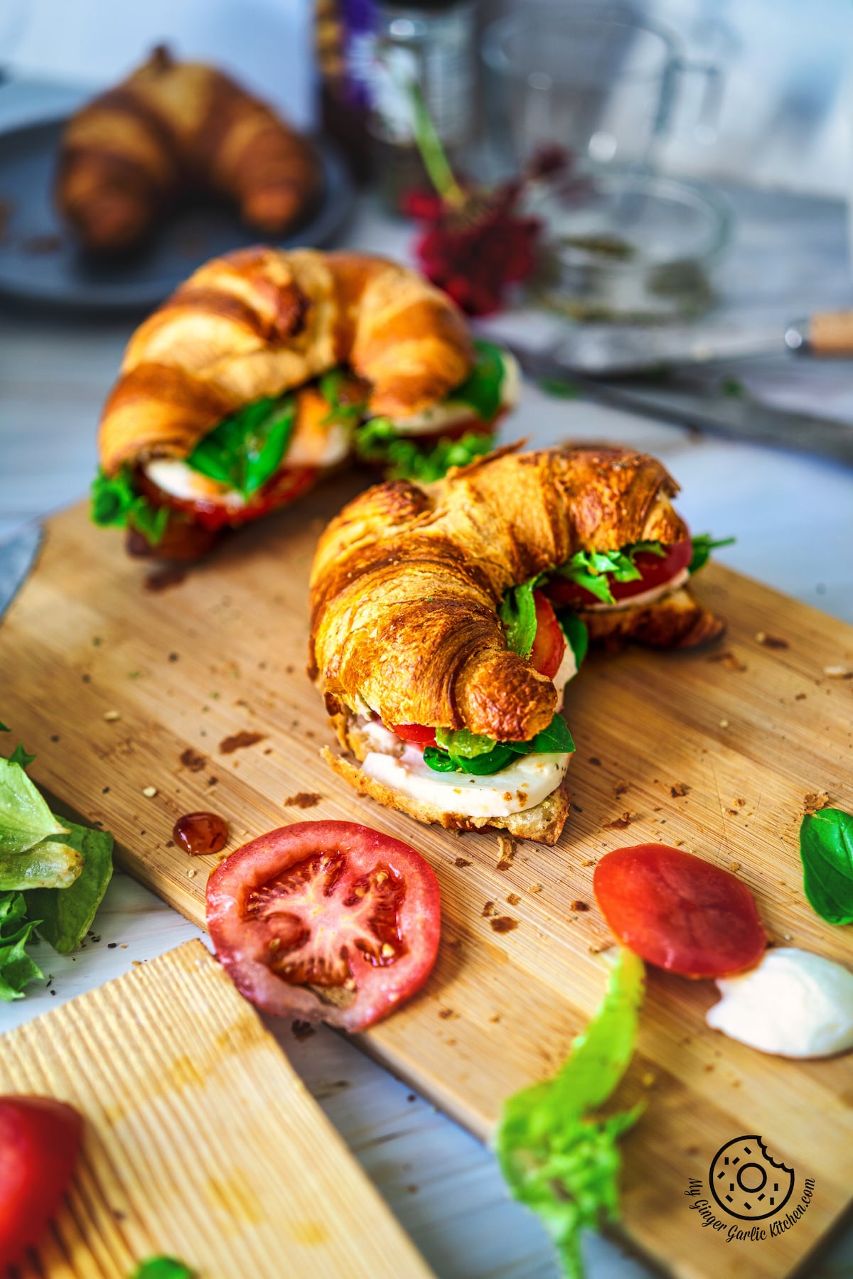 2 caprese croissant sandwices on a wooden board with tomato slice on the side