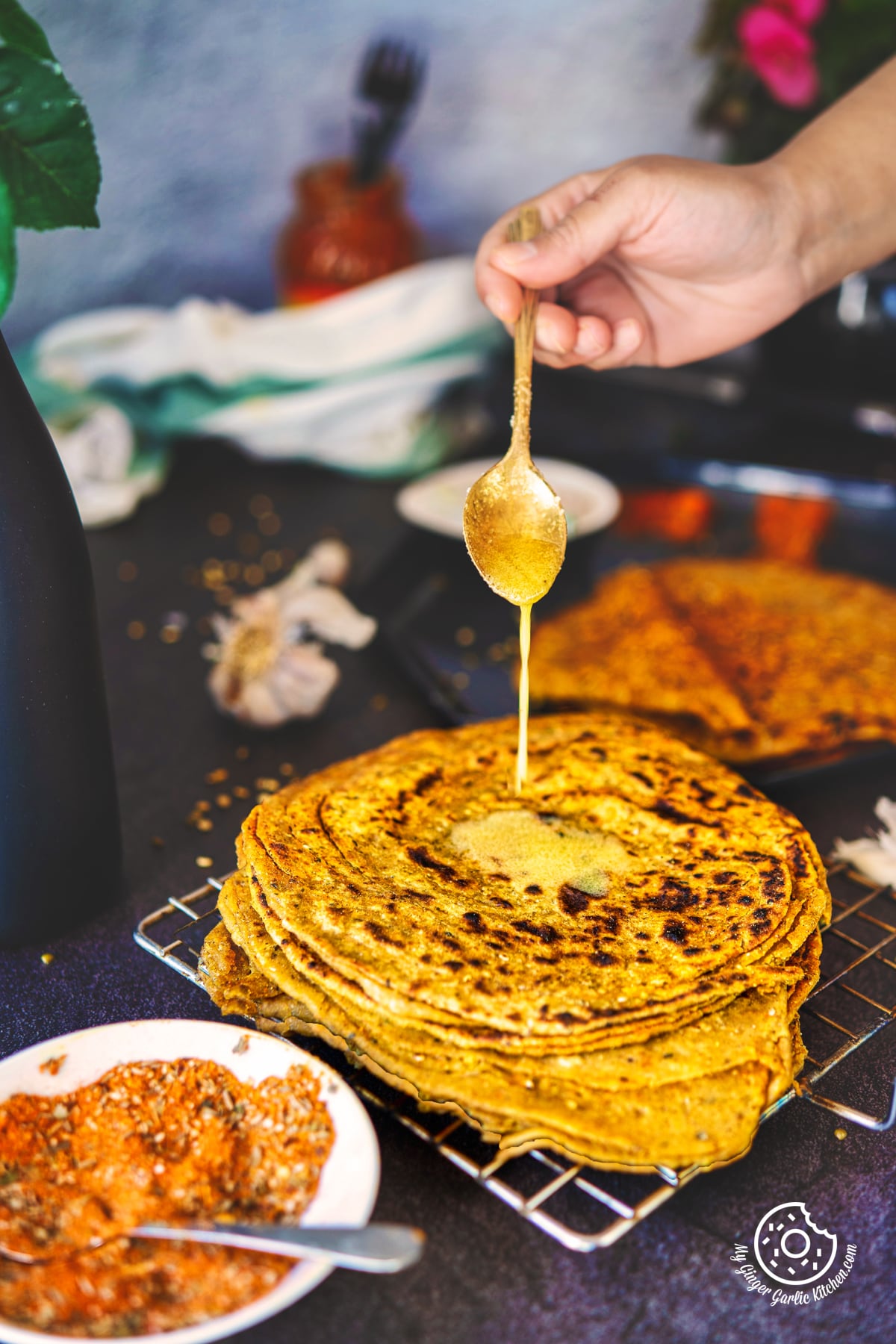 a hand drizzling ghee with a spoon over khichdi lachha paratha stacked on a wired rack