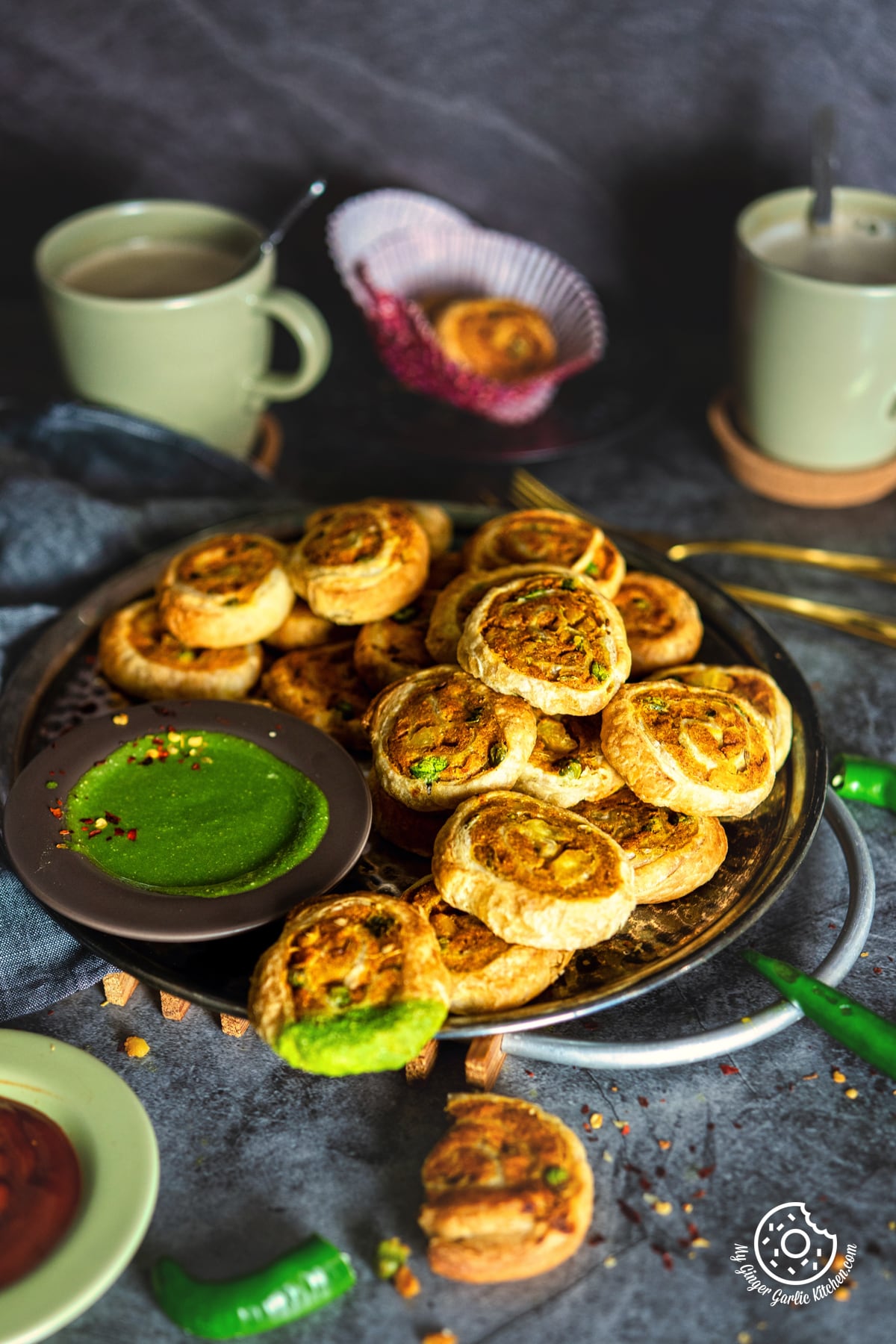 samosa pinwheels stacked on a metal plate with green coriander chutney and coffee cups in background