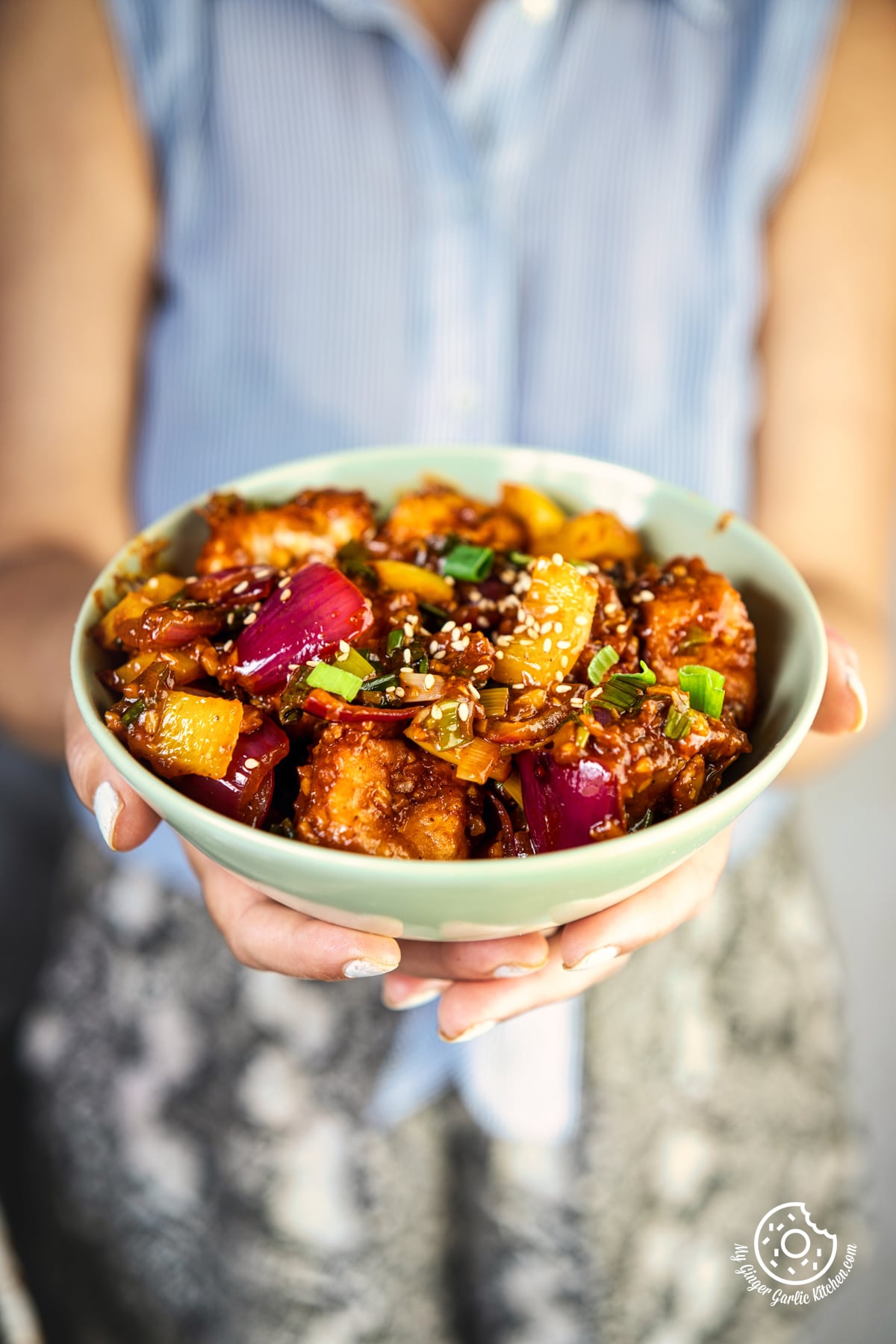 closeup of a woman holding a pale green bowl of crisp paneer manchurian topped with sesame seeds and spring greens