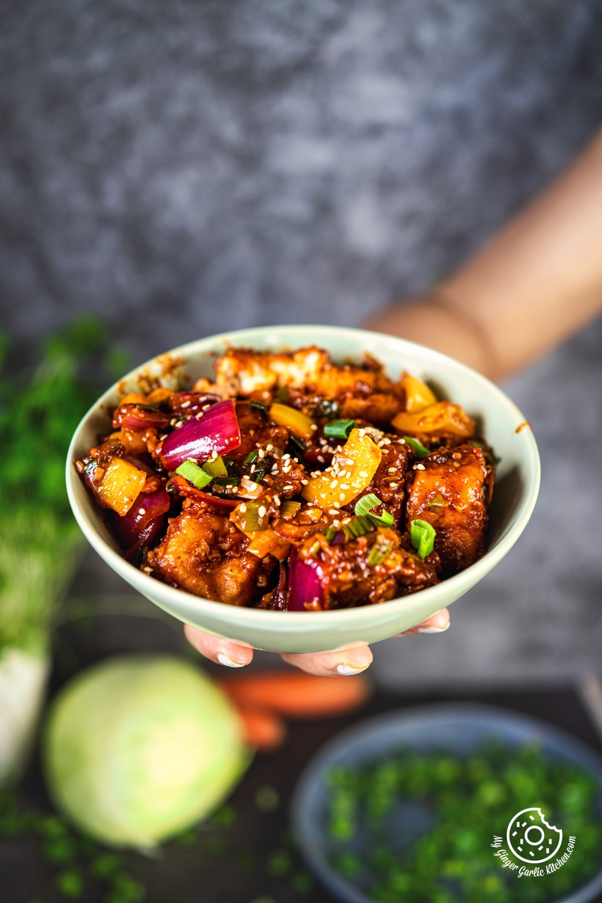 a hand holding a bowl of fried paneer manchurian topped with sesame seeds and spring greens