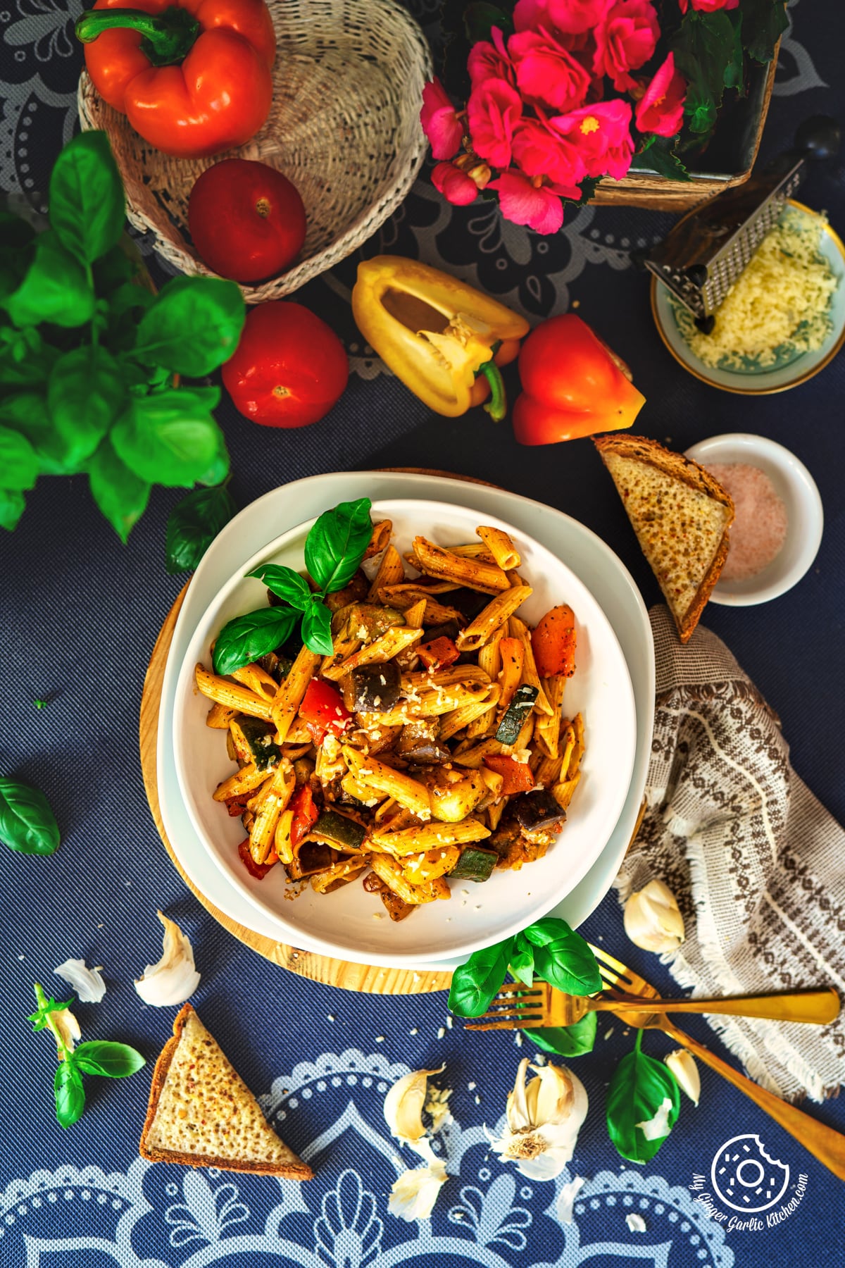 overhead shot of ratatouille pasta garnished with cheese and basil sprig served in a white plate with some fresh bell peppers and bread slices on the side
