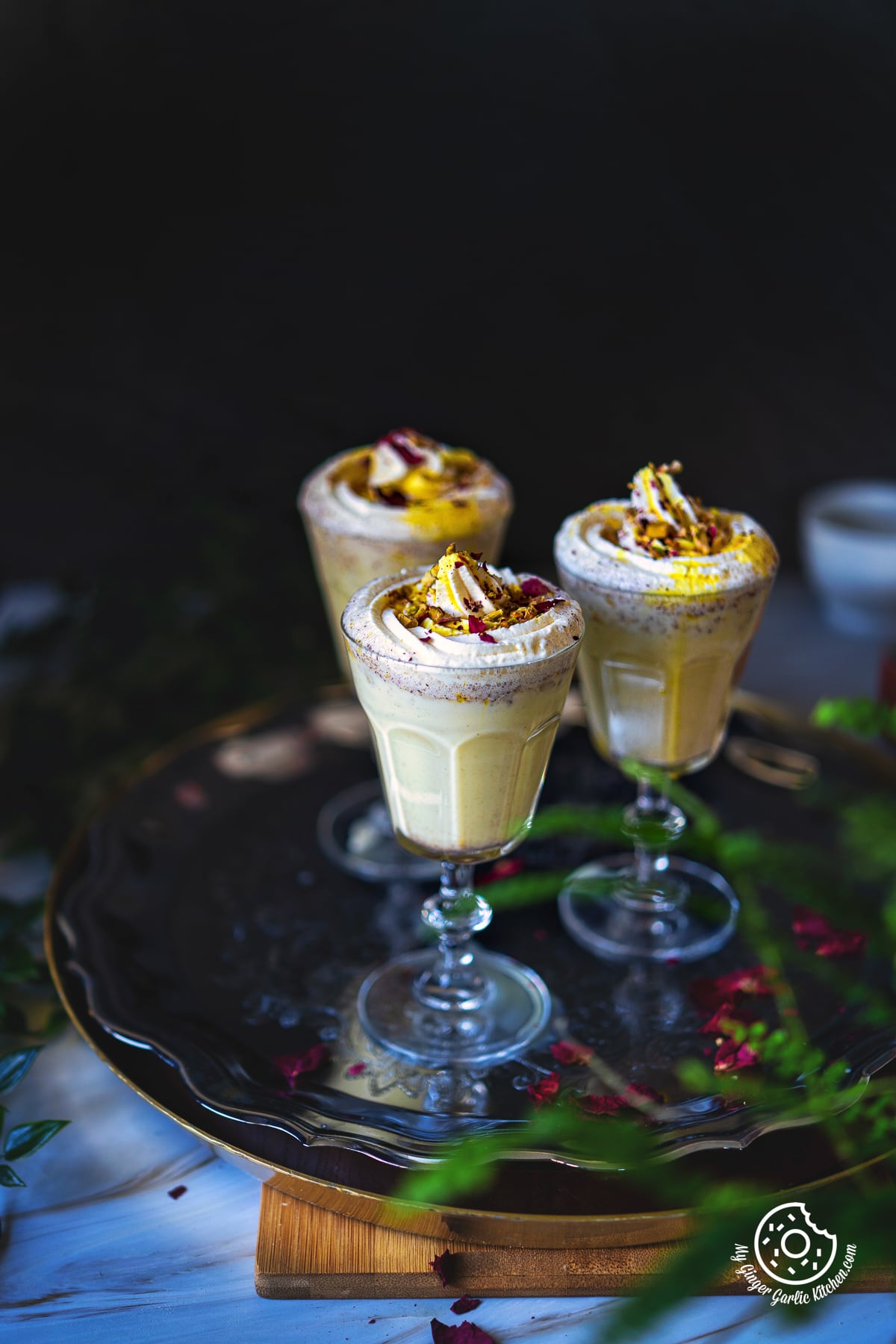 3 glasses of thandai frappe garnished with whipped cream and rose petals on a metal plate