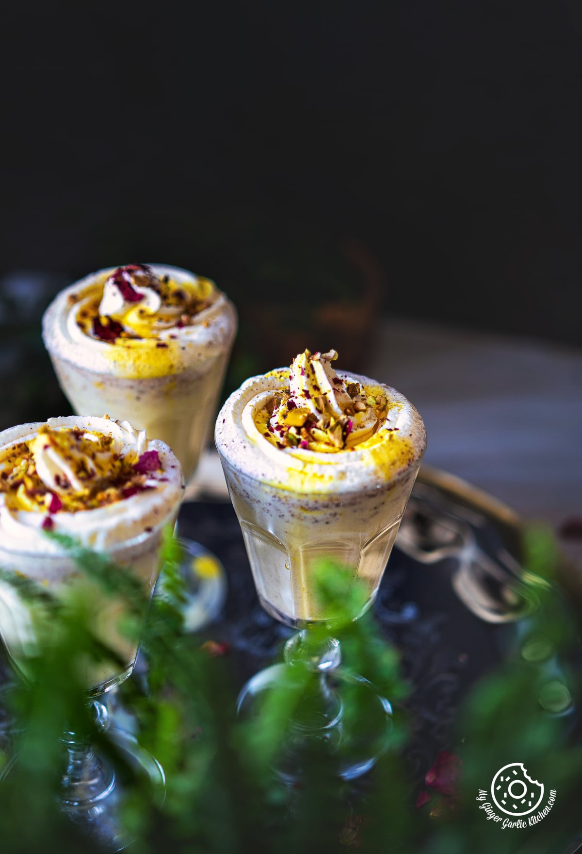 closeup of 3 glasses of thandai frappe garnished with whipped cream and rose petals on a metal plate