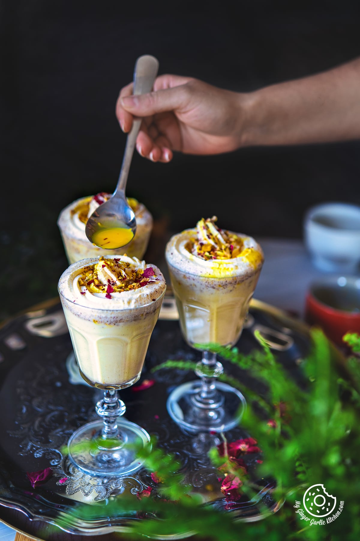 a hand holding a spoon with saffron milk over a glass of thandai frappe with teo more glasses on side