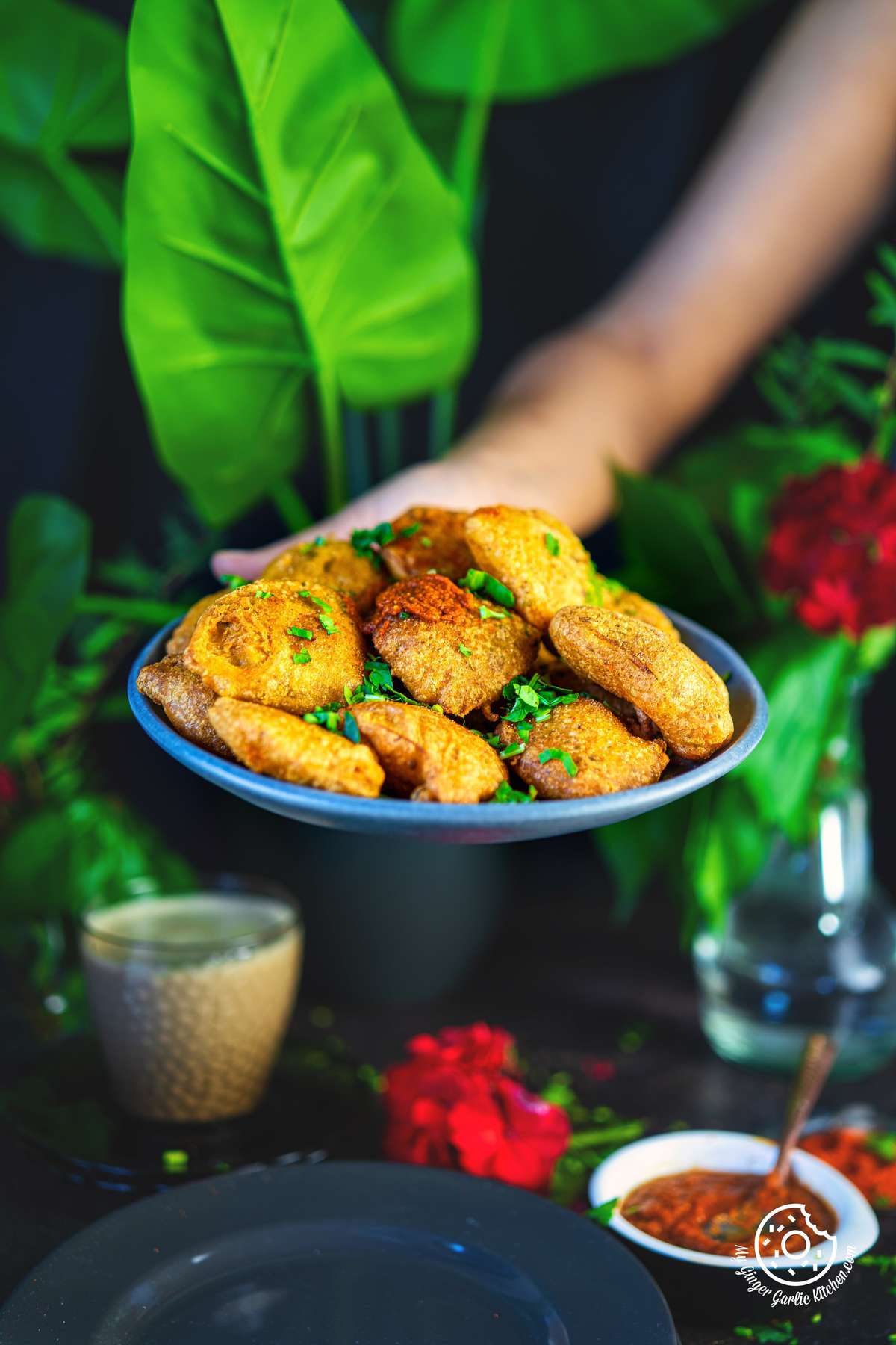 there is a person holding a plate of aloo pakora  with sauce with a glass of red flowers behind it