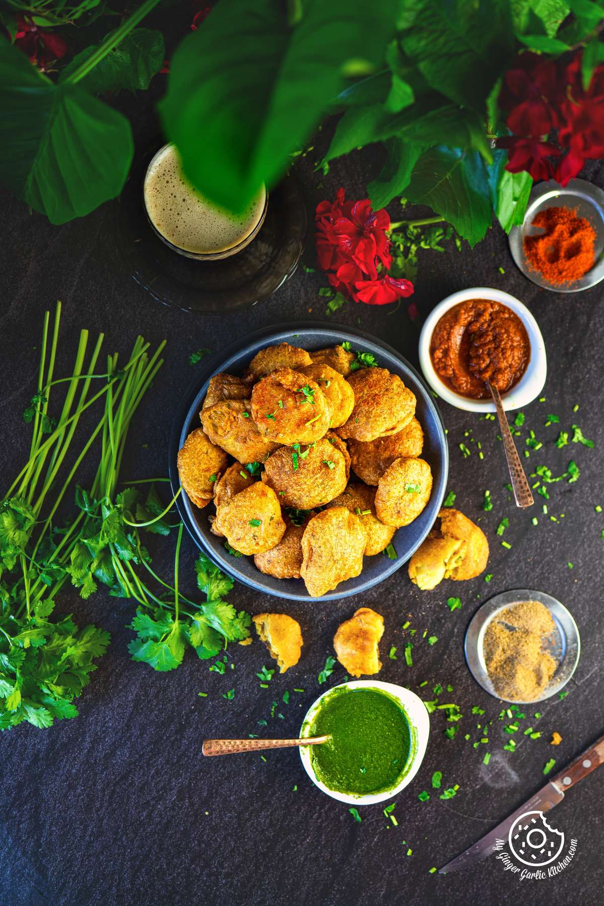 overhead photo of a a table with a bowl aloo pakoras with two chutney bowls and spices on the side
