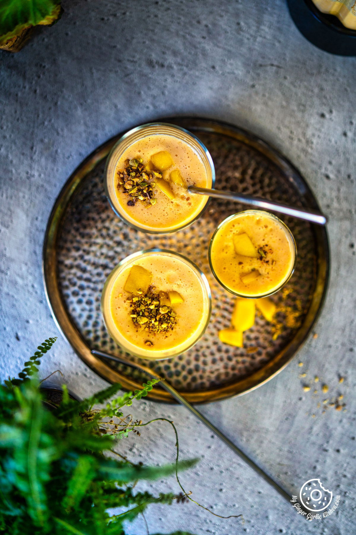 three overhead mango lassi glasses with chopped mangoes and pistachios with a plant on side