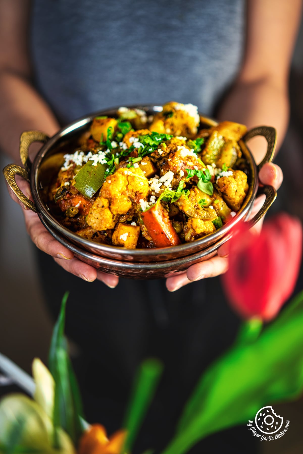 closeup of a female holding mix vegetable garnished with coriander and grated paneer in a copper kadai