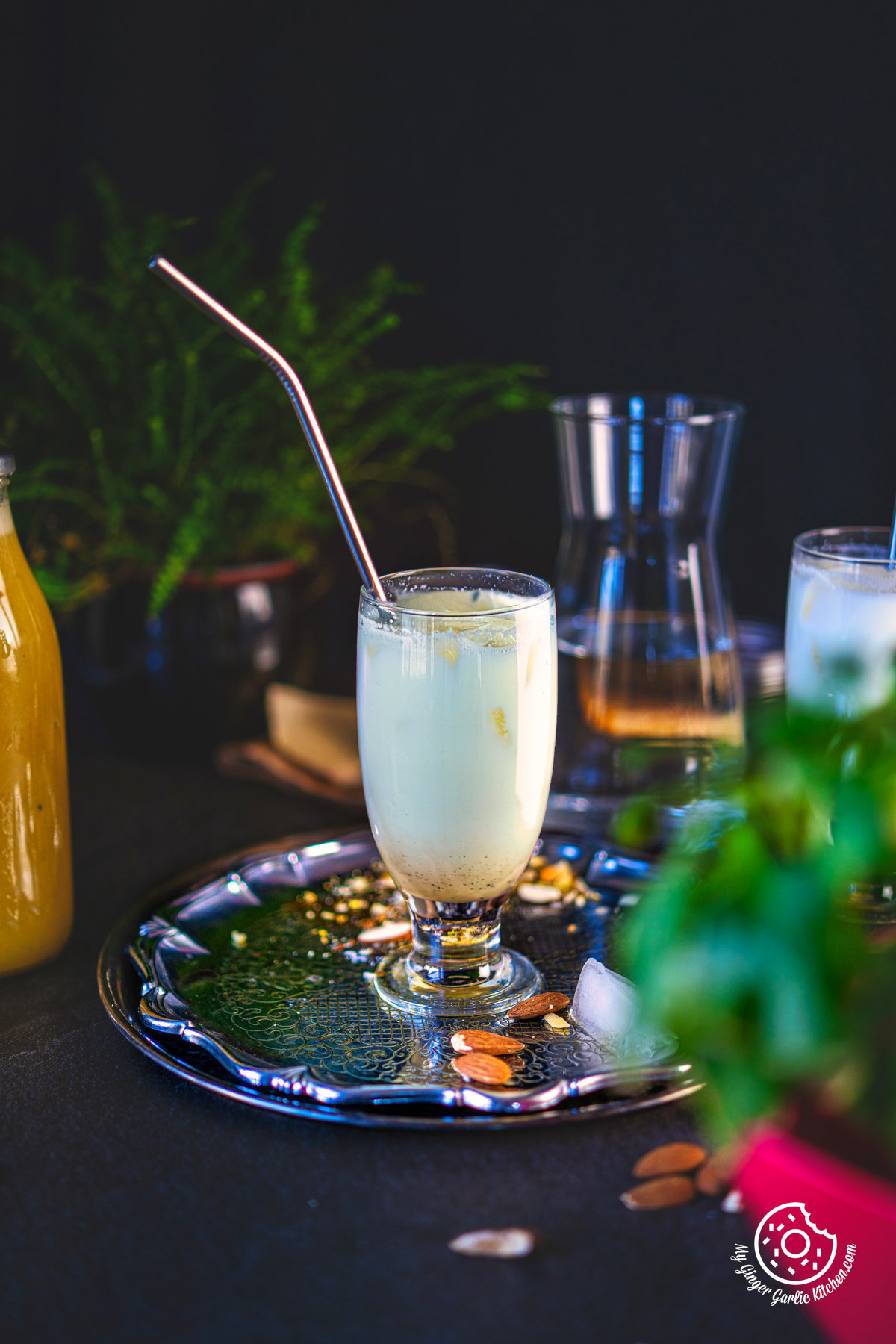 photo of a glass of badam ka sharbat on a tray with a straw and a plant in the background