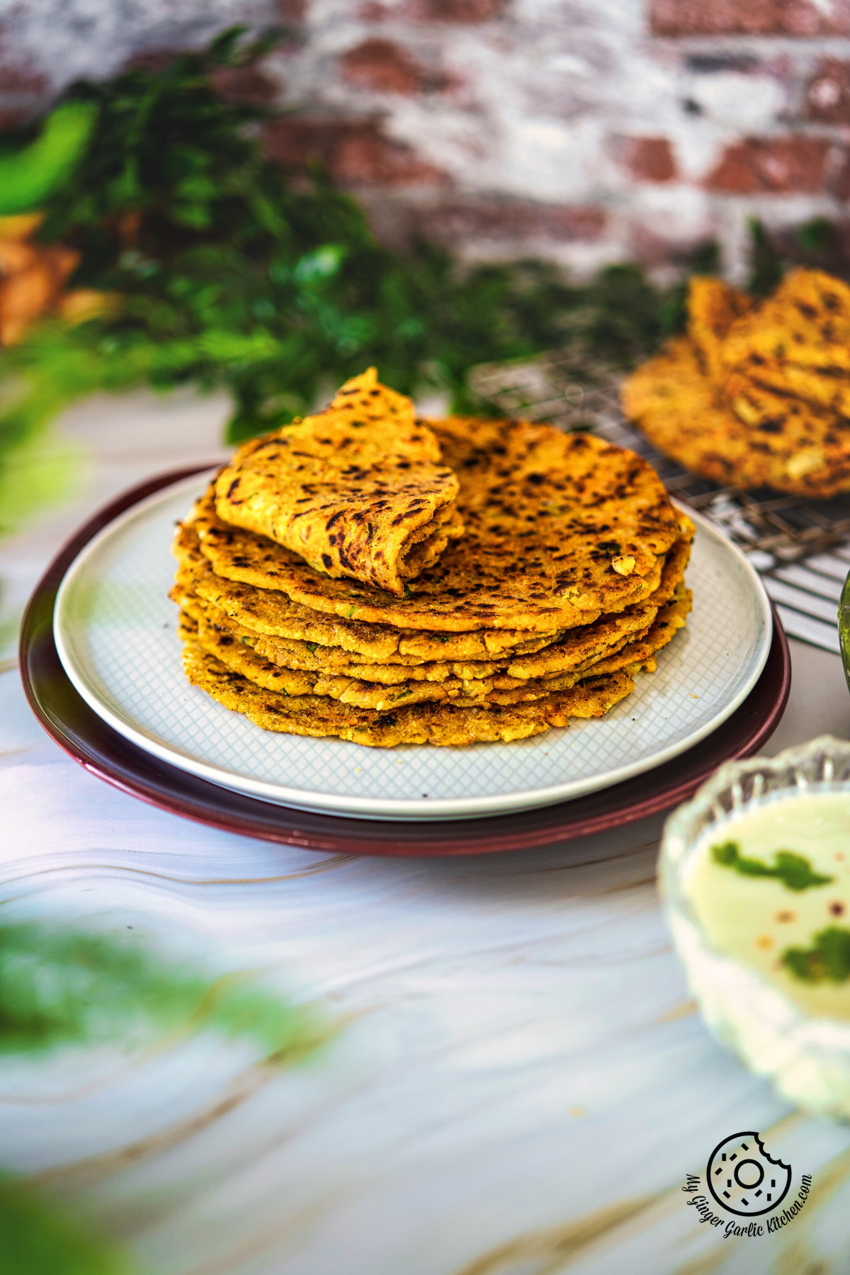 photo of a stack of rice thepla flatbreads on a plate with a bowl of raita
