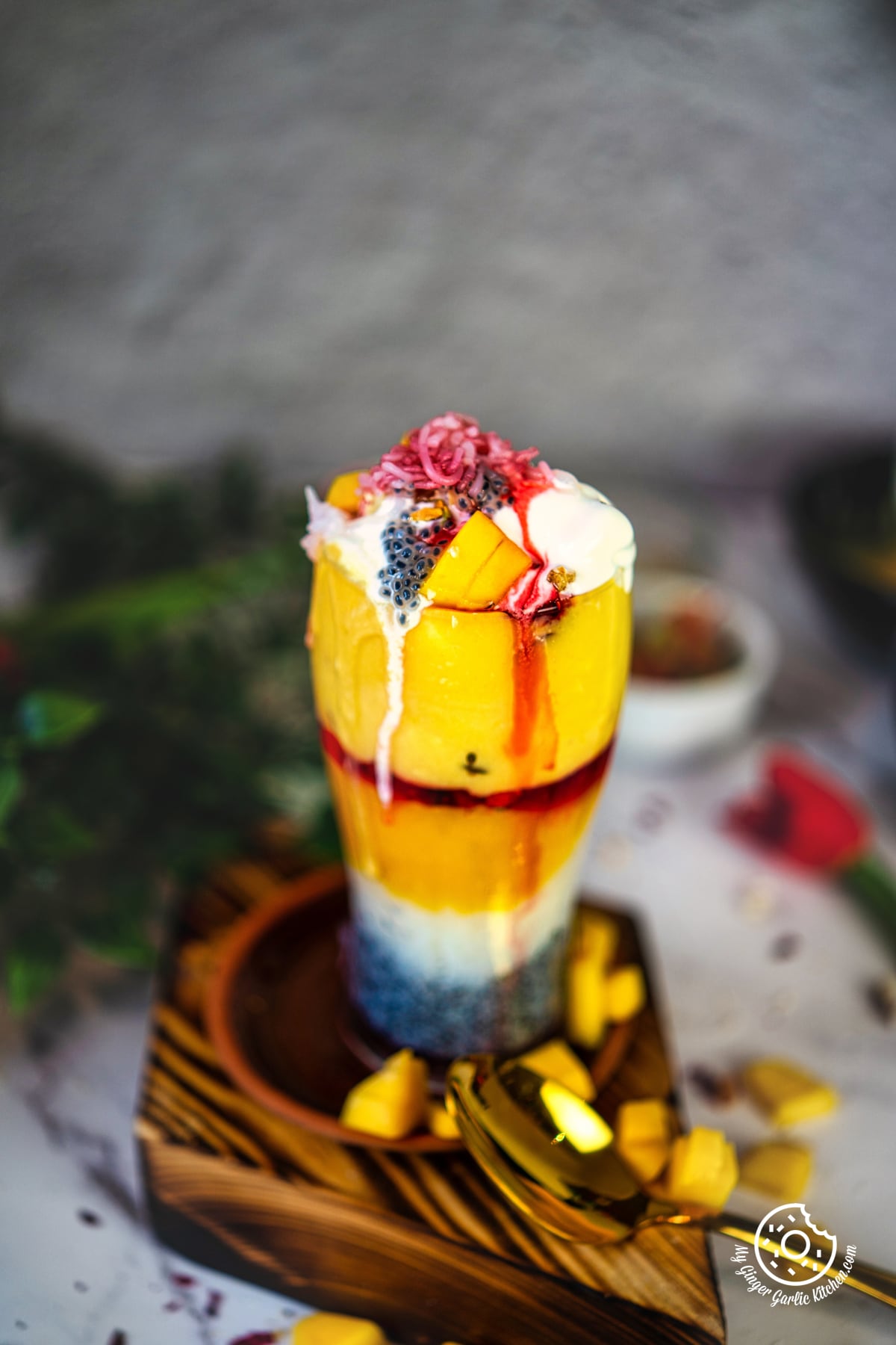 photo of a mango falooda dessert garnished with fruits and ice cream on a wooden tray with a golden spoon on the side