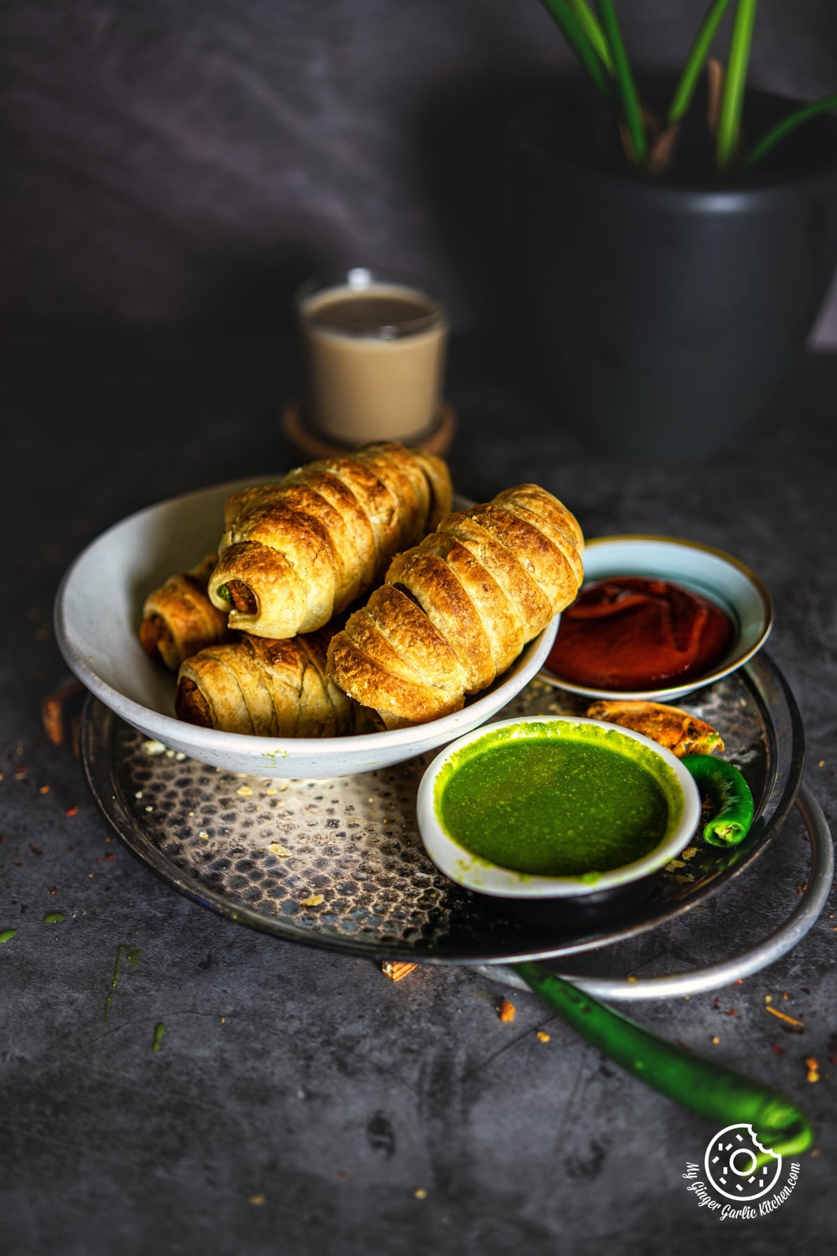a white bowl of samosa rolls kept on a metal plate with a cup of green sauce on it and a green chili on it