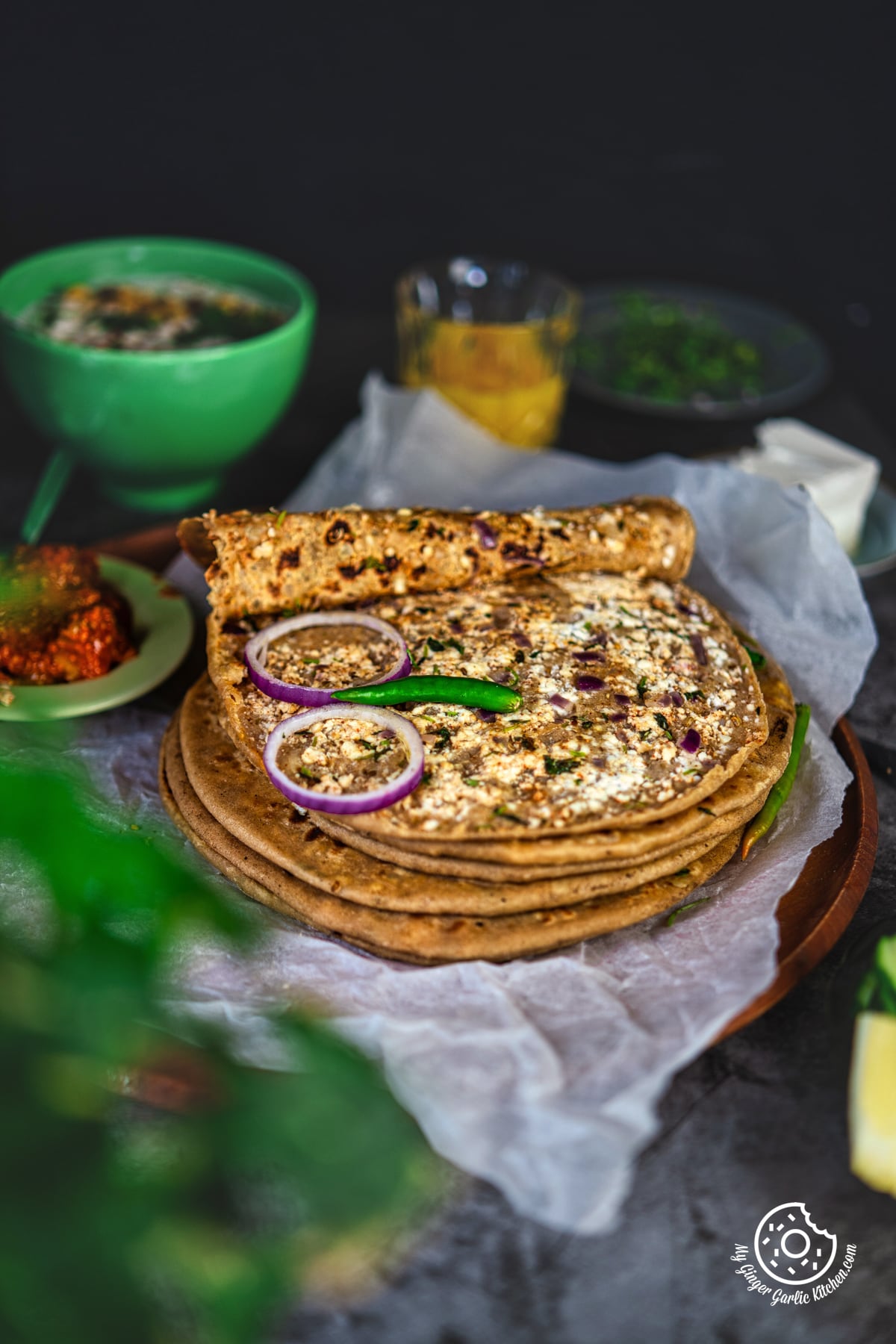 photo of paneer paratha stack on a on a wooden plate with raita and pickle in the background 